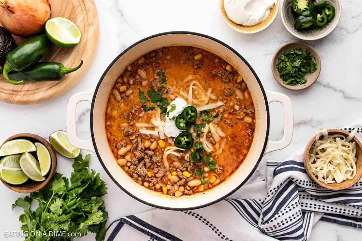 A pot of creamy taco soup reminiscent of White Bean Turkey Chili, topped with shredded cheese, sliced jalapeños, sour cream, and cilantro. Surrounding the pot are bowls of chopped cilantro, lime wedges, fresh vegetables, and more cheese.