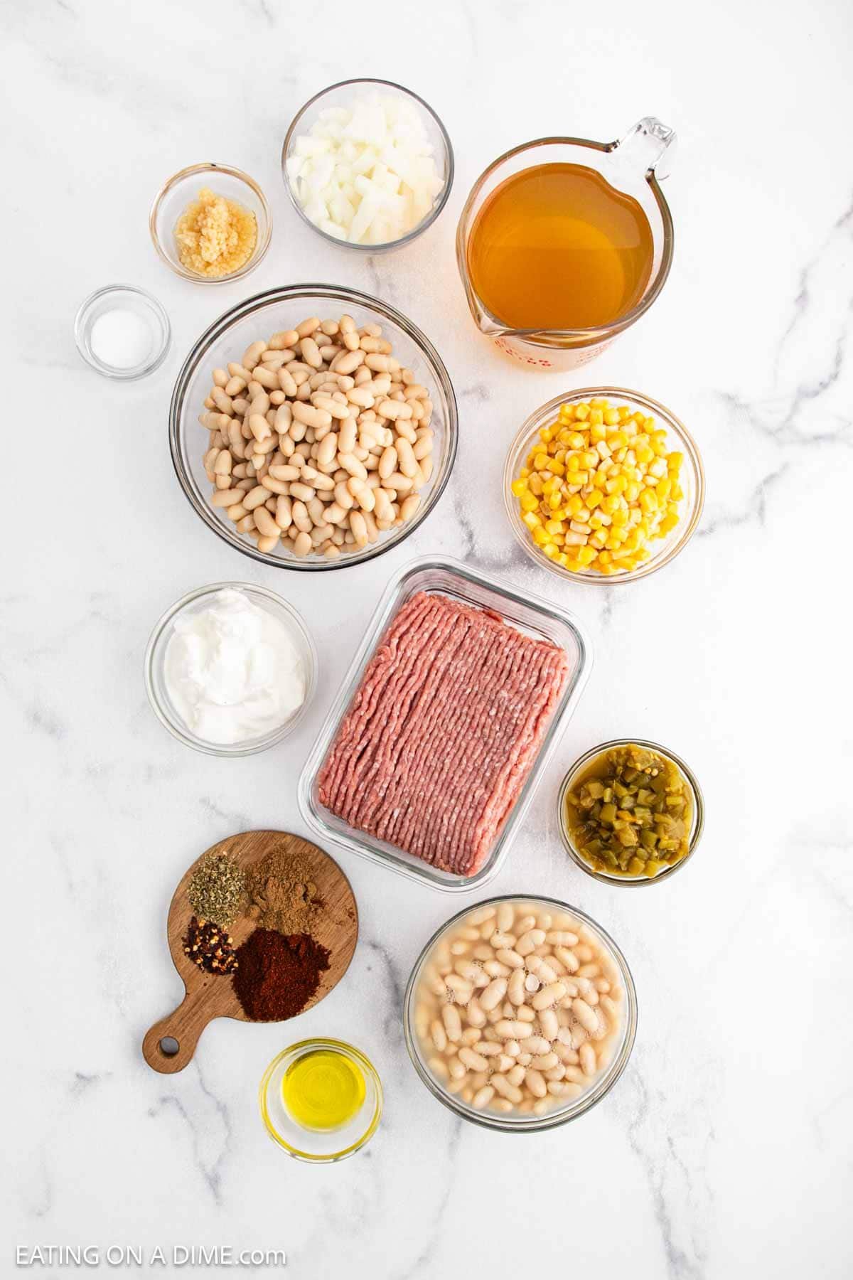 Various ingredients for a White Bean Turkey Chili are arranged on a white surface, including ground turkey, canned white beans, corn, chopped onions, chicken broth, diced green chilies, seasonings, oil, garlic, and sour cream.
