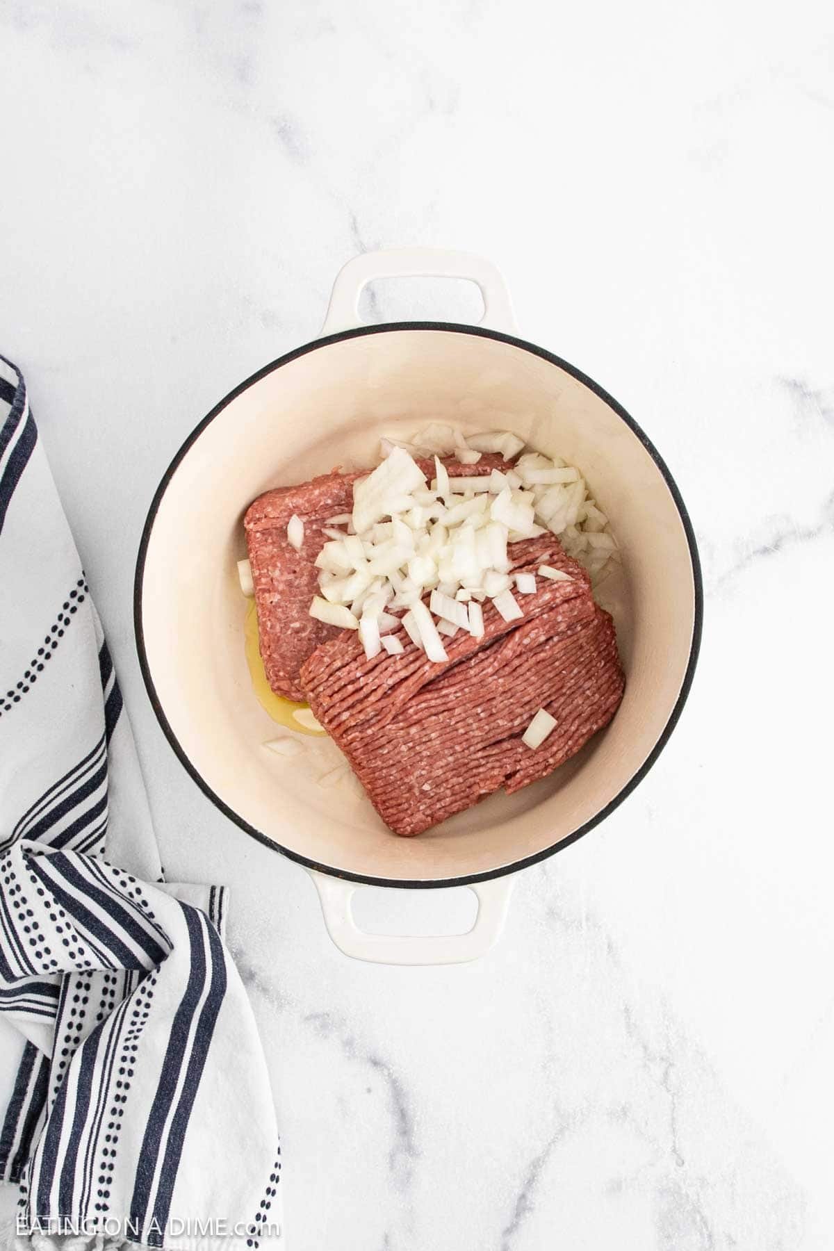 A white pot on a marble surface holds raw ground beef and chopped onions, ready for chili. A black and white striped kitchen towel is positioned to the left of the pot.
