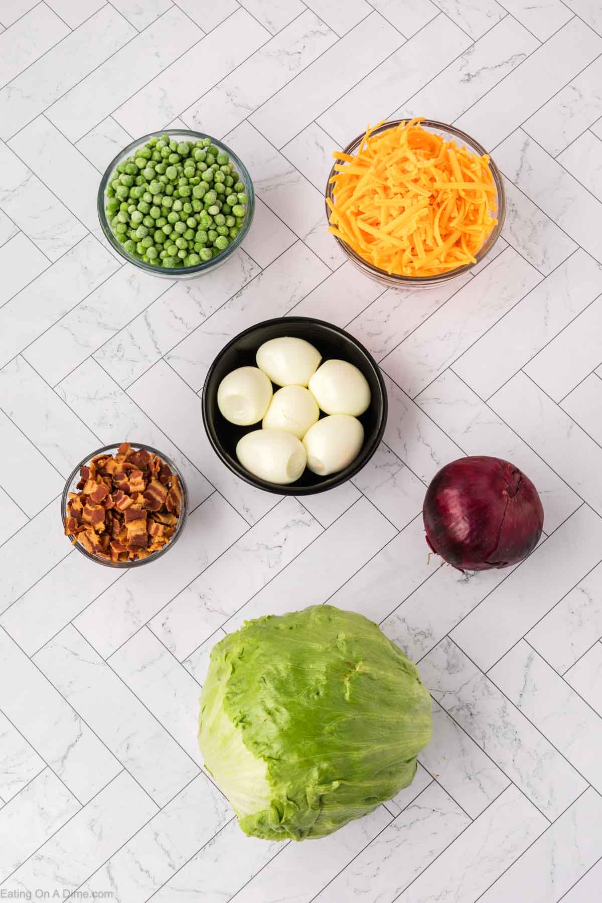 Six ingredients for a classic layered salad are arranged on a white tile surface: shredded cheddar cheese, green peas, sliced hard-boiled eggs, diced cooked bacon, a red onion, and a head of iceberg lettuce. Perfect for any 7 Layer Salad recipe.