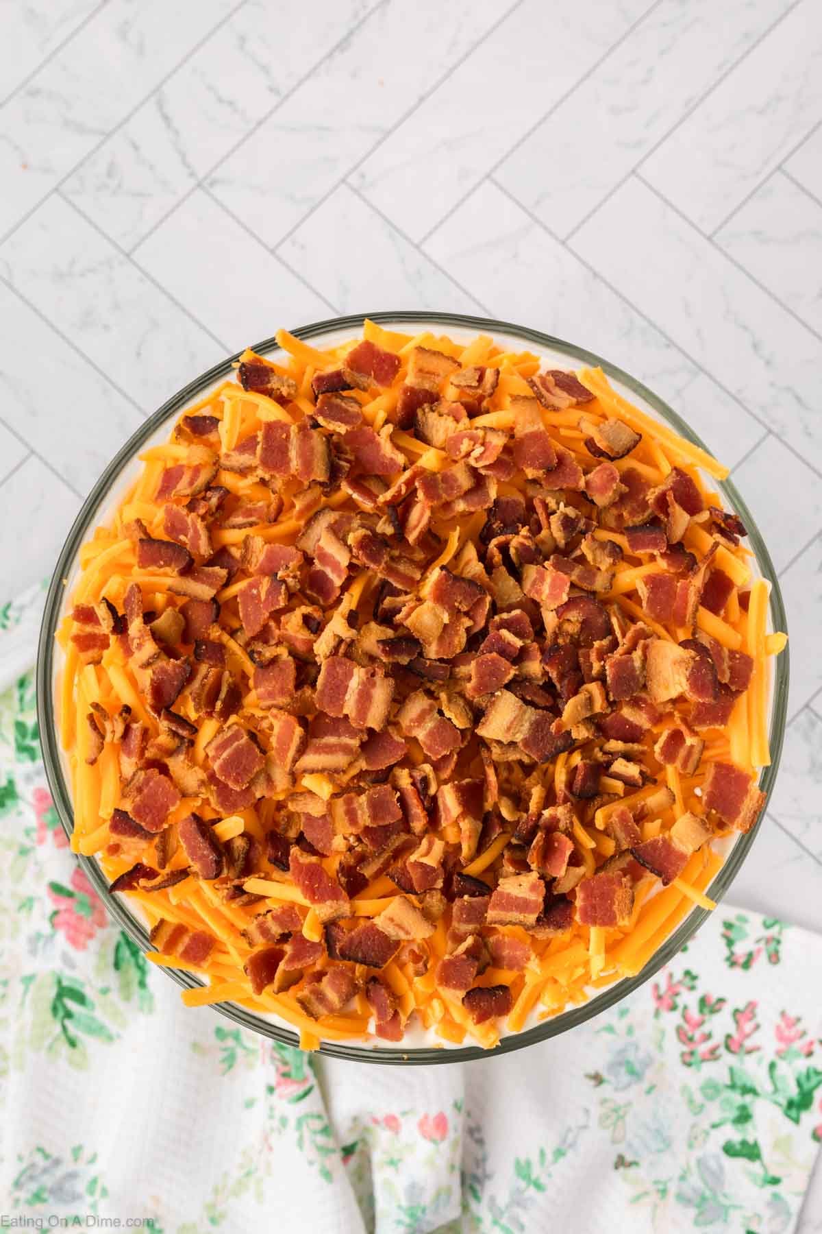 A glass bowl filled with 7 Layer Salad, topped with shredded cheddar cheese and crispy bacon pieces, placed on a white, floral-patterned cloth with a tiled background.