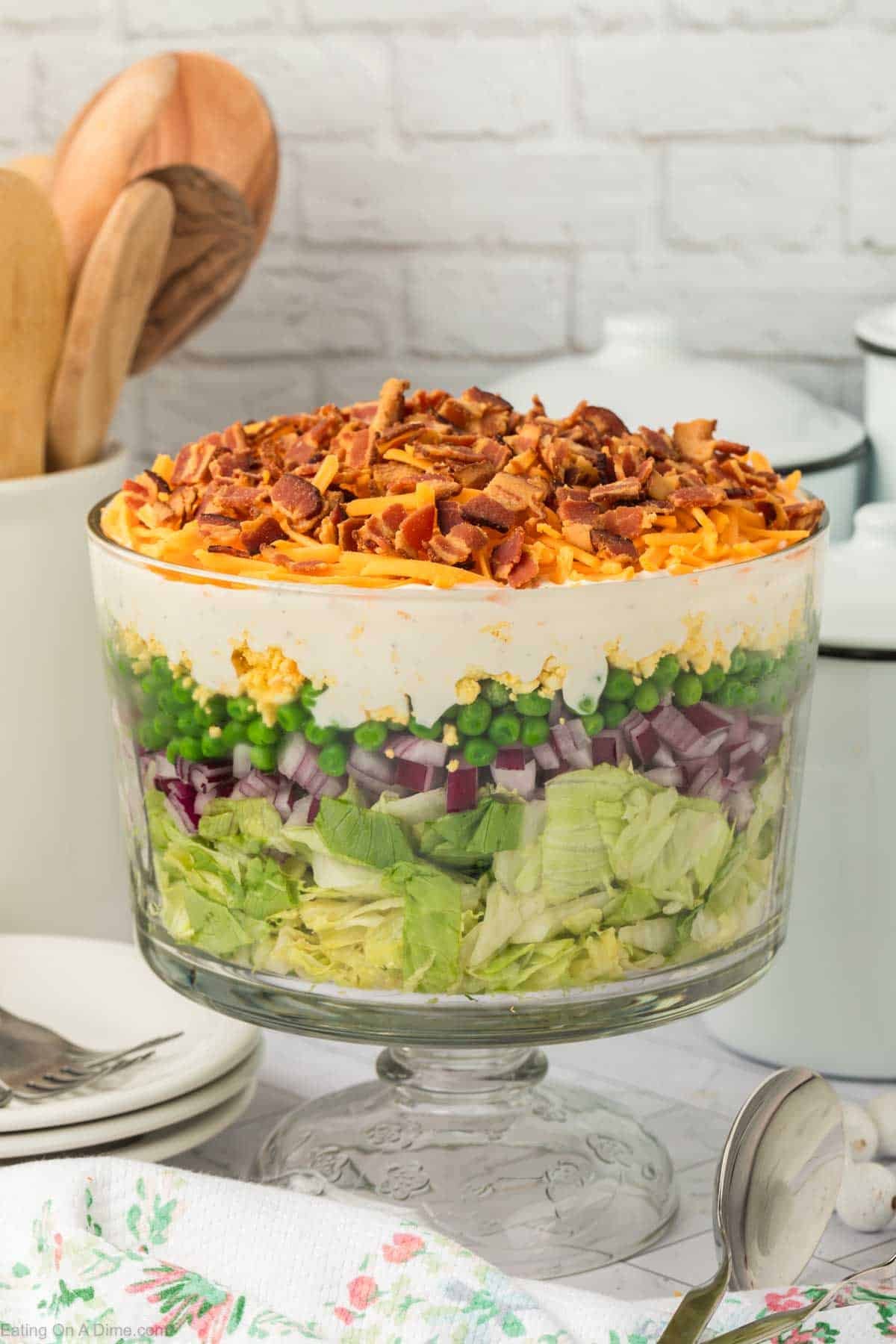A glass trifle bowl filled with classic 7 layer salad—crisp lettuce, red onions, peas, shredded cheese, a creamy layer, and crumbled bacon—sits on a table set with plates and utensils.