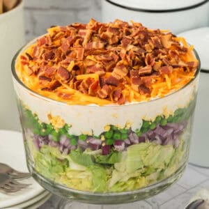 A clear glass bowl filled with classic 7 Layer Salad, featuring chopped lettuce, red onion, green peas, creamy white dressing, shredded cheddar cheese, and crumbled bacon layered beautifully on top.