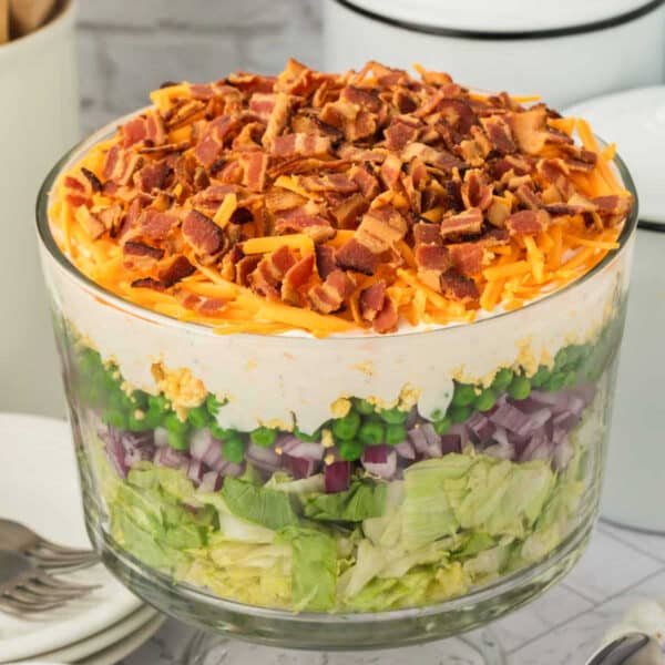 A clear glass bowl filled with classic 7 Layer Salad, featuring chopped lettuce, red onion, green peas, creamy white dressing, shredded cheddar cheese, and crumbled bacon layered beautifully on top.