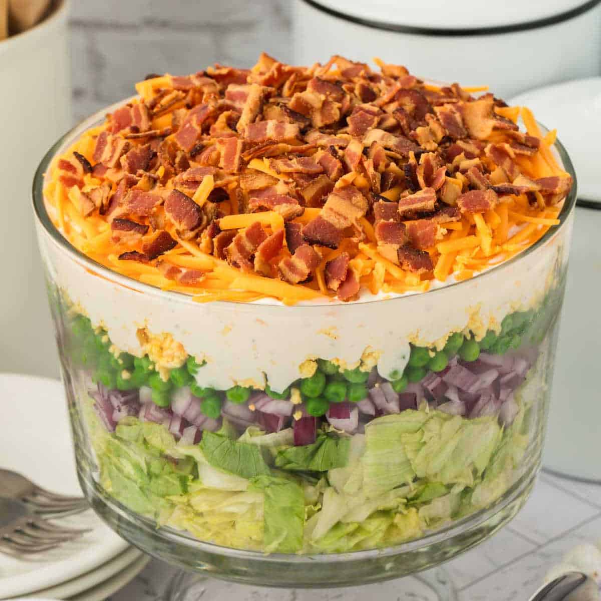 A clear glass bowl filled with classic 7 Layer Salad, featuring chopped lettuce, red onion, green peas, creamy white dressing, shredded cheddar cheese, and crumbled bacon layered beautifully on top.