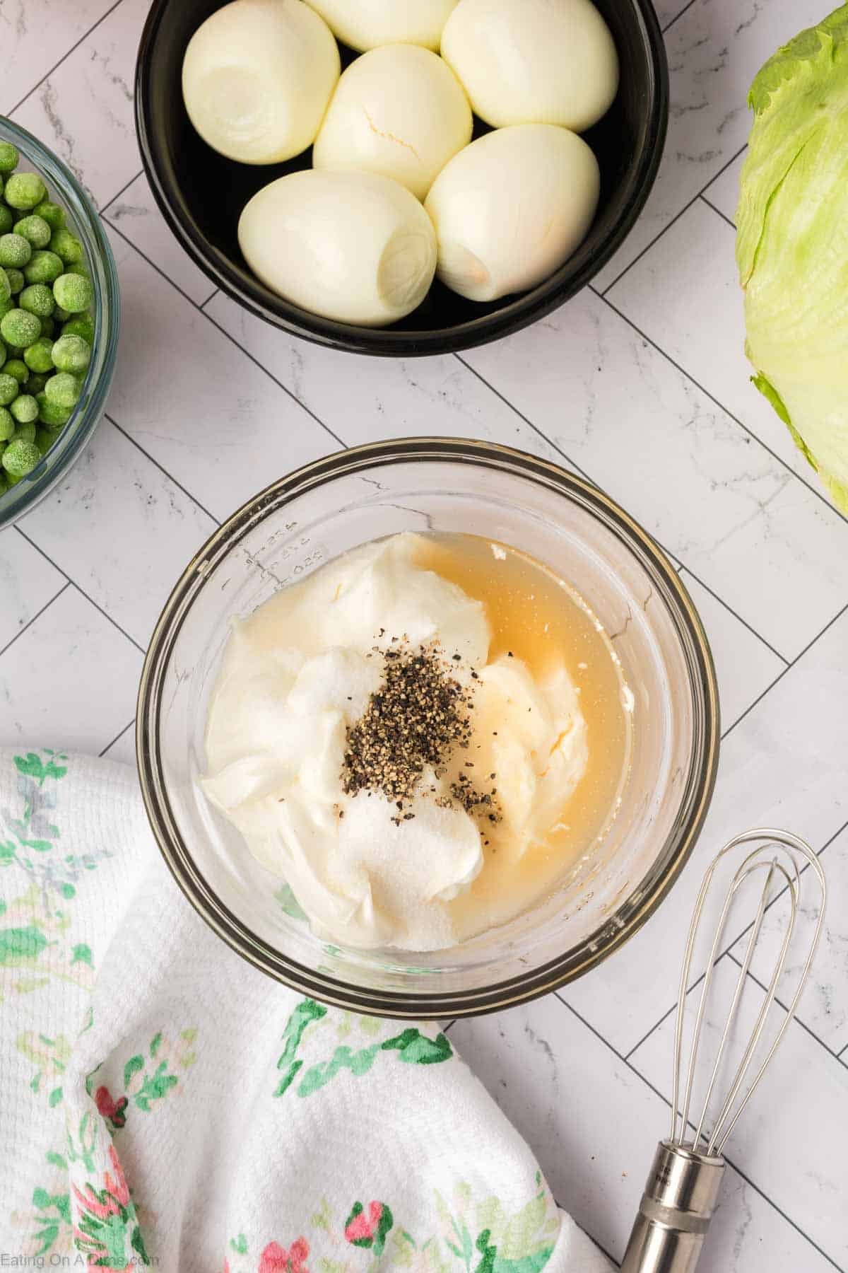 A bowl with mayonnaise, vinegar, sugar, and black pepper—classic 7 Layer Salad ingredients—sits on a countertop next to a whisk, hard-boiled eggs, green peas, a head of lettuce, and a floral towel.