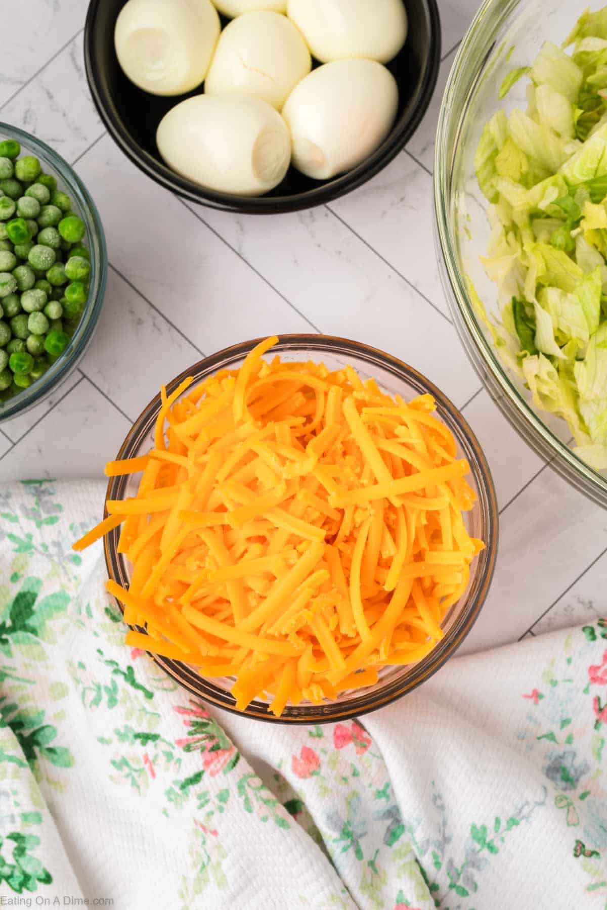 A bowl of shredded cheddar cheese sits on a table near bowls of green peas, hard-boiled eggs, and chopped lettuce—all classic ingredients for a 7 Layer Salad—with a floral-patterned cloth nearby.