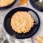 A serving of creamy, cheesy corn casserole sits on a dark blue plate, surrounded by forks, shredded cheese, and a patterned napkin on a white tiled surface—the perfect cheesy corn recipe for any occasion.