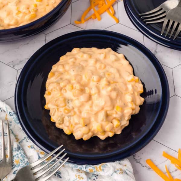 A serving of creamy, cheesy corn casserole sits on a dark blue plate, surrounded by forks, shredded cheese, and a patterned napkin on a white tiled surface—the perfect cheesy corn recipe for any occasion.