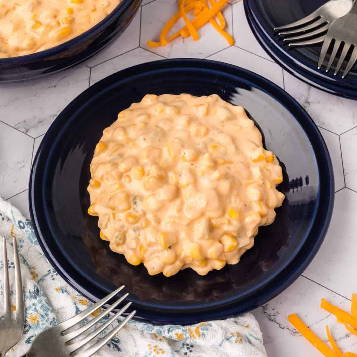 A serving of creamy, cheesy corn casserole sits on a dark blue plate, surrounded by forks, shredded cheese, and a patterned napkin on a white tiled surface—the perfect cheesy corn recipe for any occasion.