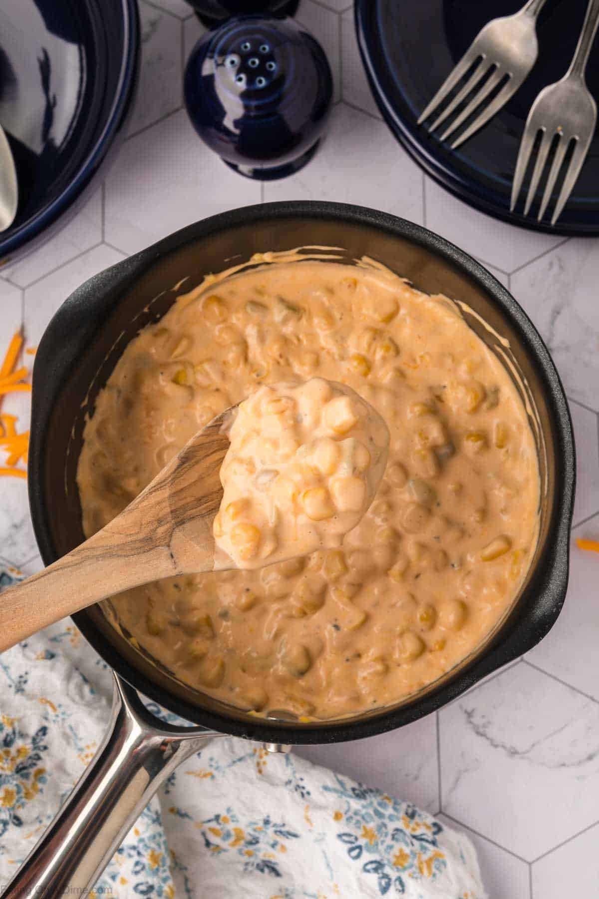 A saucepan filled with creamy, cheesy corn casserole—a delicious take on a classic corn recipe—being stirred with a wooden spoon, surrounded by blue plates, forks, a salt shaker, and a floral kitchen towel on a white tiled surface.