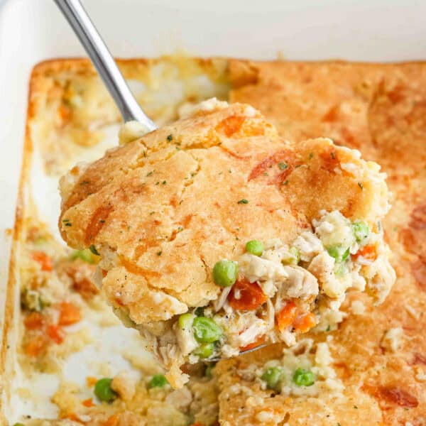 A serving of chicken cobbler with a golden biscuit topping is being lifted from a baking dish, revealing peas, carrots, and chicken in a creamy filling.