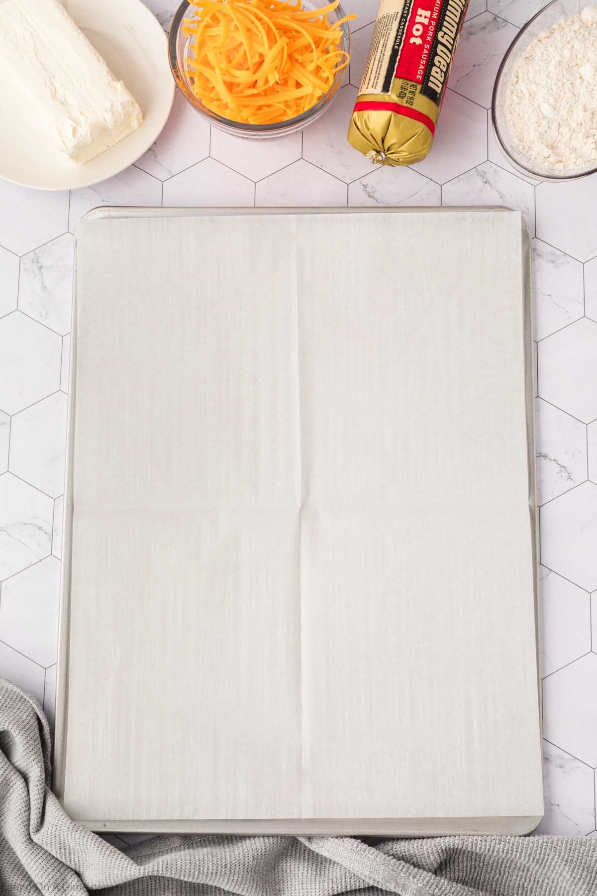 A lined baking sheet on a white hexagonal tile surface, surrounded by all the ingredients you need to make cream cheese sausage balls—shredded cheddar, a block of cream cheese, sausage, flour, and a gray cloth.