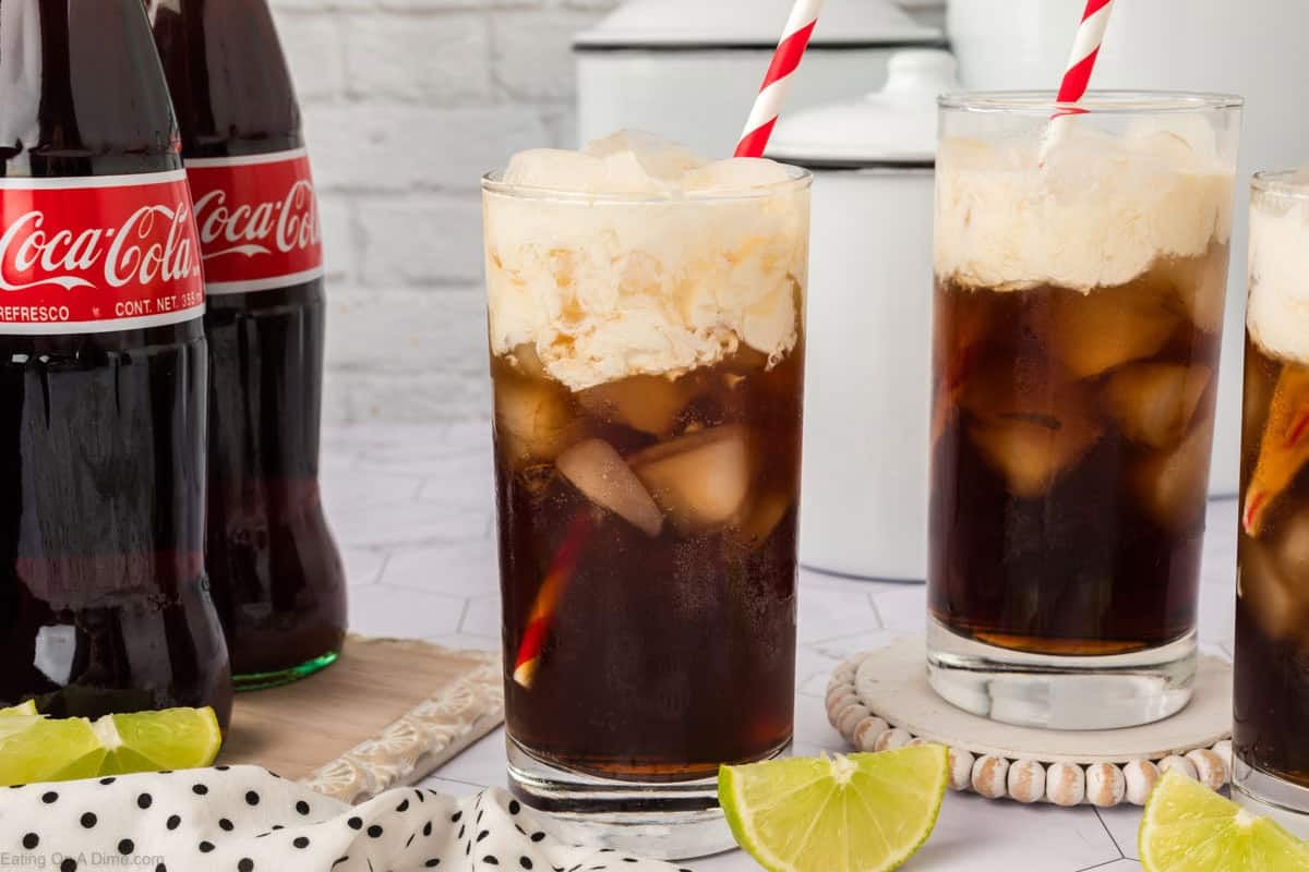 Three glasses of cola with ice and vanilla ice cream float, crafted as a classic Dirty Soda Recipe, each with a red and white striped straw, sit beside glass Coca-Cola bottles, lime wedges, and a white cloth with black dots.