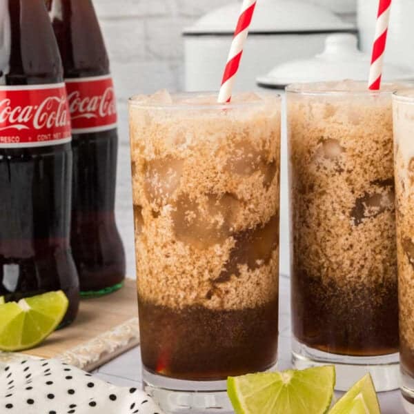 Three tall glasses of ice cream floats with frothy tops and red-striped straws are in the foreground, surrounded by lime wedges, showcasing a fun dirty soda recipe. Coca-Cola bottles and a polka dot cloth appear in the background.