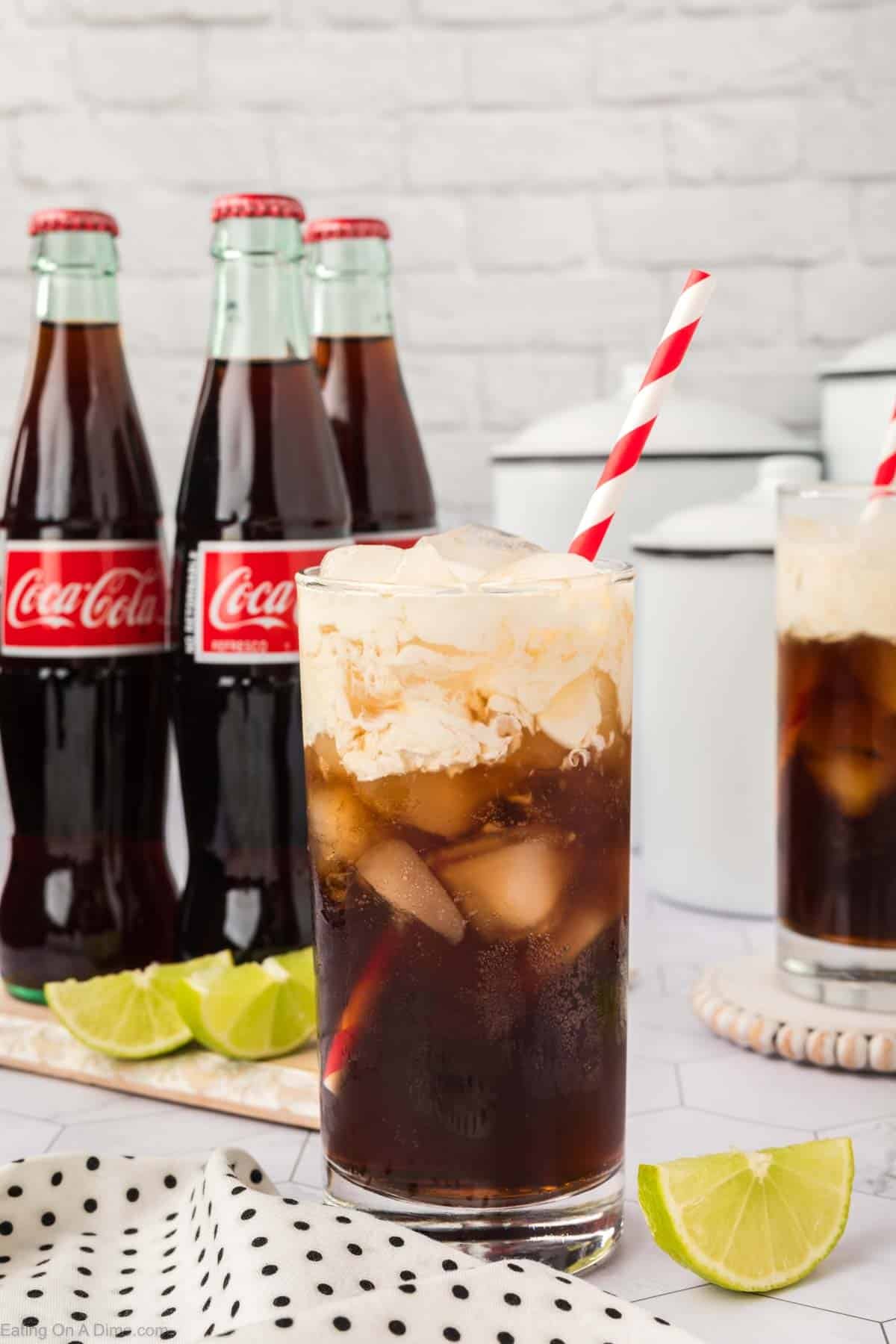 A glass of cola with ice cream float and a red-striped straw sits on a polka dot cloth, featuring a fun Dirty Soda Recipe vibe, with lime wedges and classic glass Coca-Cola bottles in the background.