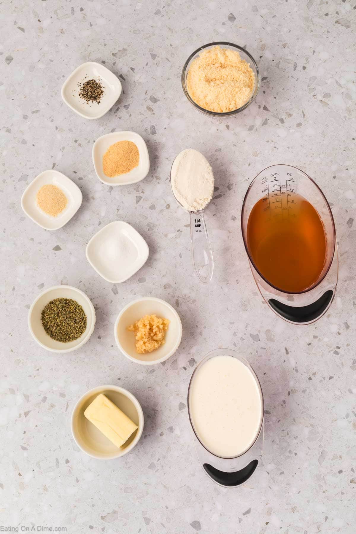 Ingredients for Garlic Parmesan Sauce are displayed on a gray countertop, with butter, heavy cream, broth, grated cheese, flour, minced garlic, dried herbs, pepper, salt, onion powder, and garlic powder in small bowls.