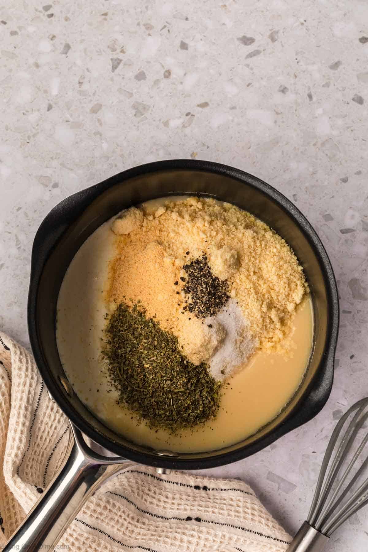 A black saucepan on a light countertop holds the beginnings of a rich Garlic Parmesan Sauce—cream, grated cheese, dried herbs, garlic powder, pepper, and salt—ready to be mixed. A whisk and beige striped towel sit beside the pan.