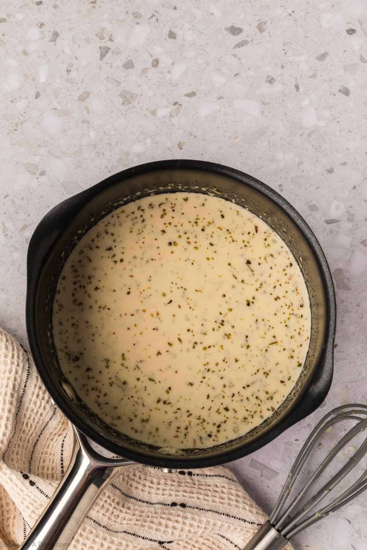 A saucepan filled with creamy Garlic Parmesan Sauce speckled with herbs, placed on a light-colored countertop next to a beige textured cloth and a metal whisk.