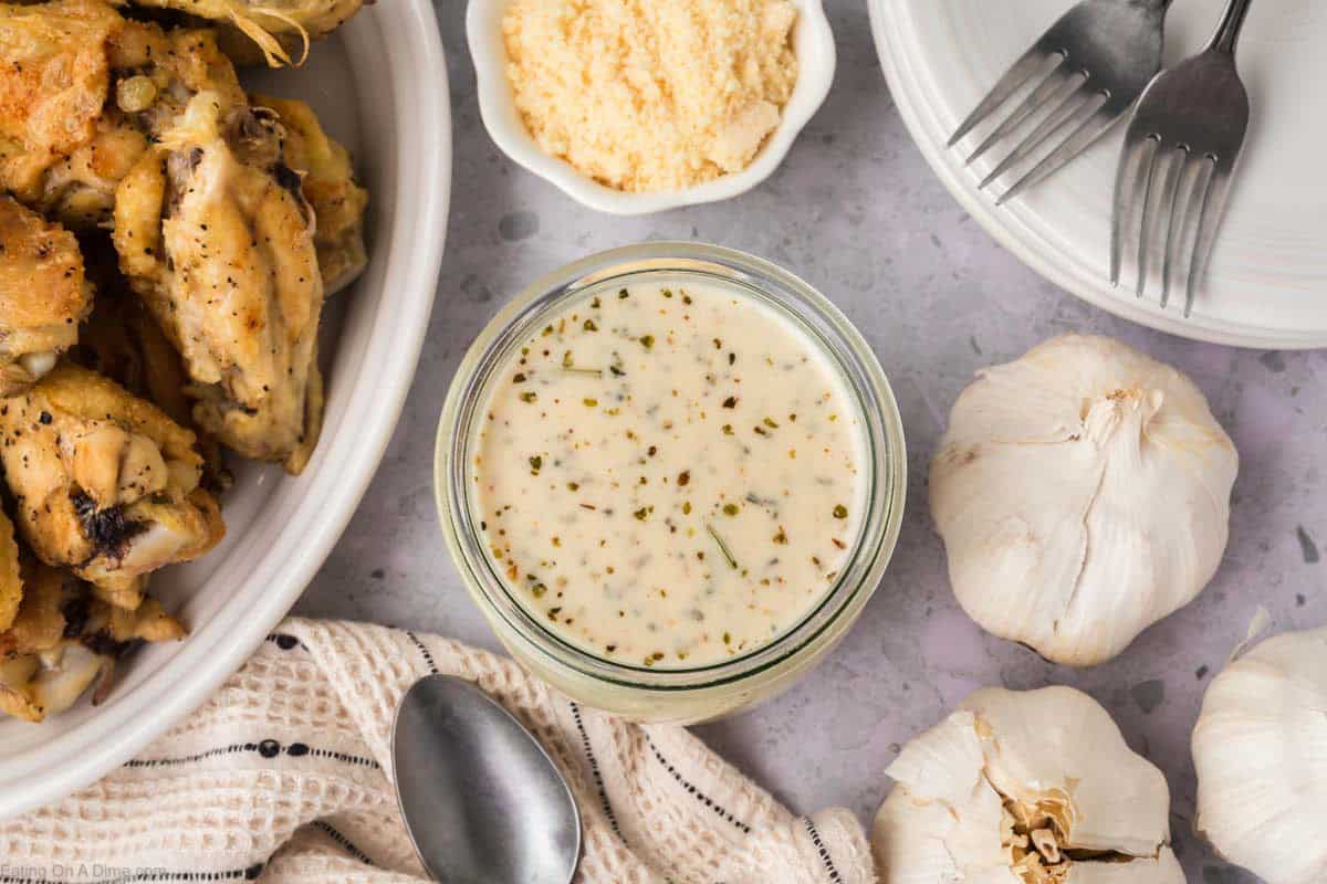 A glass jar of creamy, herb-speckled Garlic Parmesan Sauce sits on a gray surface near garlic bulbs, a bowl of grated cheese, a plate with forks, and a dish of cooked chicken wings. A spoon and white cloth are also visible.