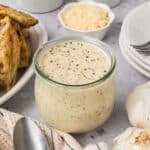 A glass jar filled with creamy Italian sauce sits on a gray countertop, surrounded by a spoon, a bulb of garlic, plates, parmesan cheese, and cooked chicken pieces.