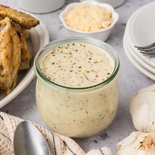 A glass jar filled with creamy Italian sauce sits on a gray countertop, surrounded by a spoon, a bulb of garlic, plates, parmesan cheese, and cooked chicken pieces.