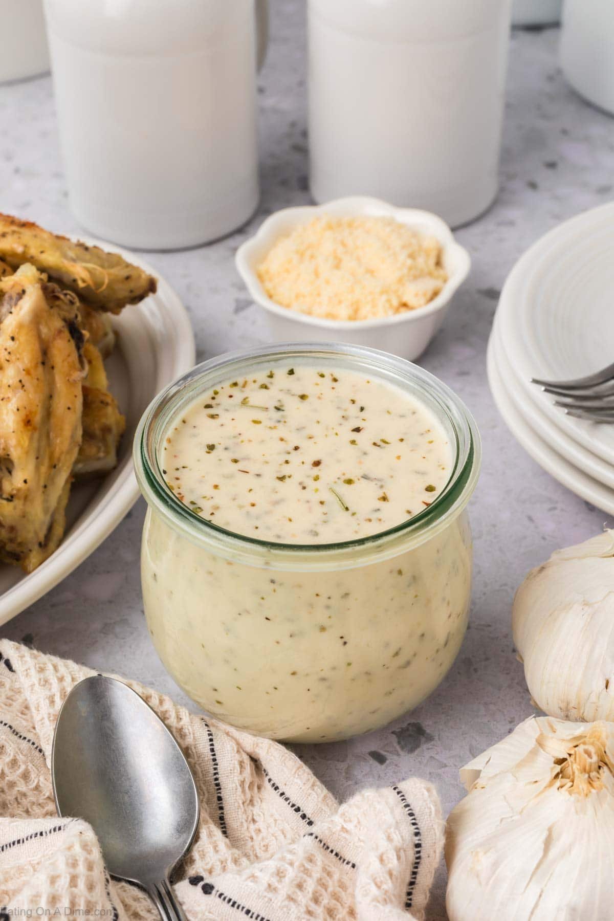 A glass jar filled with creamy, herb-speckled Garlic Parmesan Sauce sits on a countertop near a spoon, fresh garlic bulbs, grated cheese, cooked chicken wings, and stacked white plates.