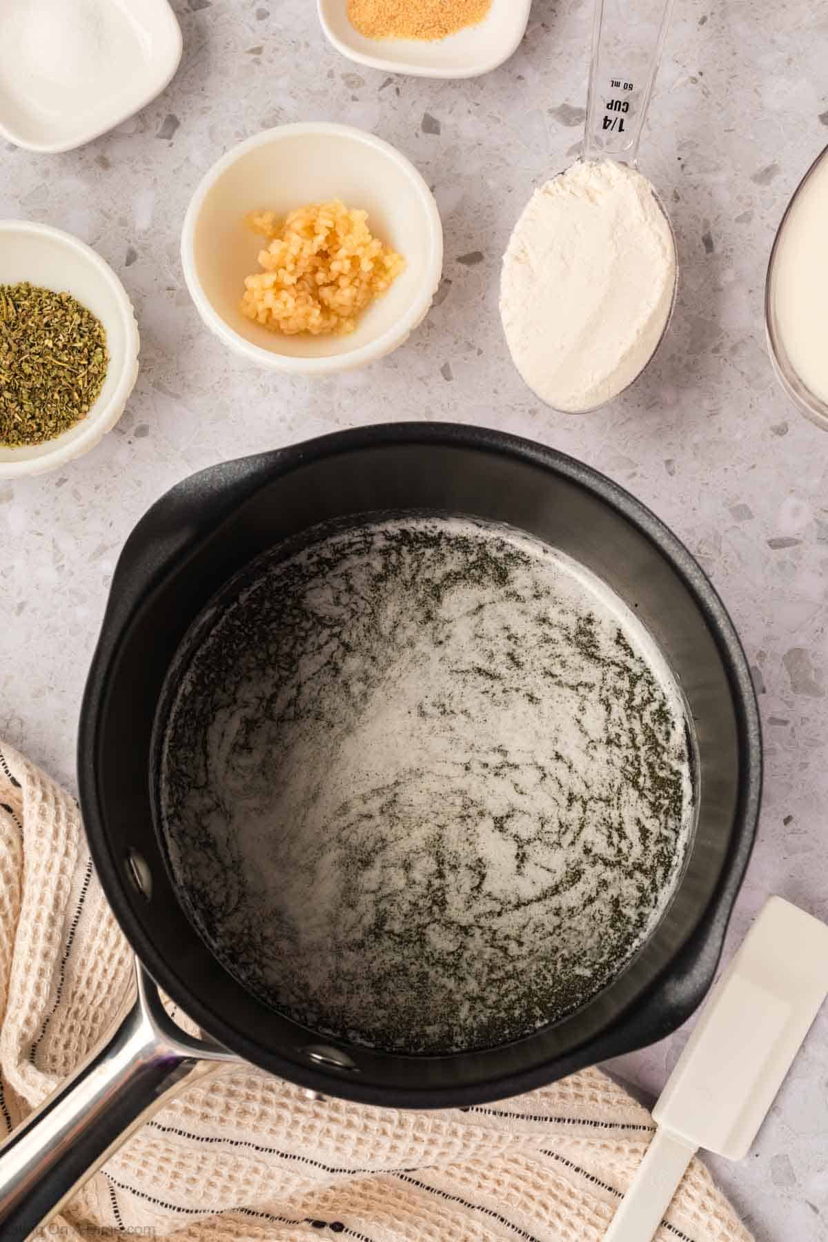A saucepan with melted butter for Garlic Parmesan Sauce sits on a countertop, surrounded by bowls of minced garlic, dried herbs, flour, and a measuring cup of milk. A spatula and kitchen towel are nearby, ready for cooking.