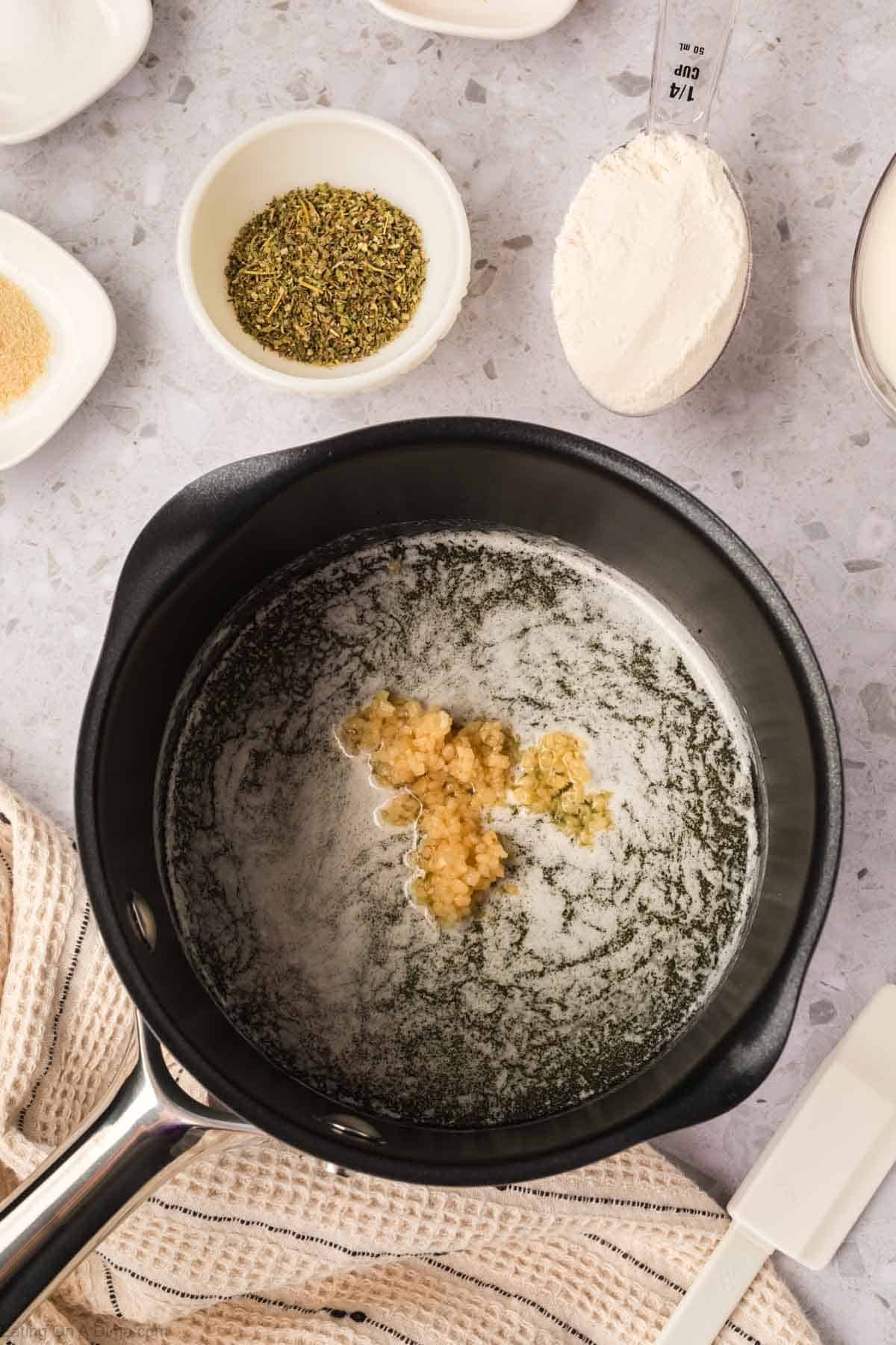A saucepan with melted butter and minced garlic inside, perfect for making Garlic Parmesan Sauce, is surrounded by small bowls of dried herbs, flour, and spices on a light countertop. A measuring spoon and kitchen towel are nearby.