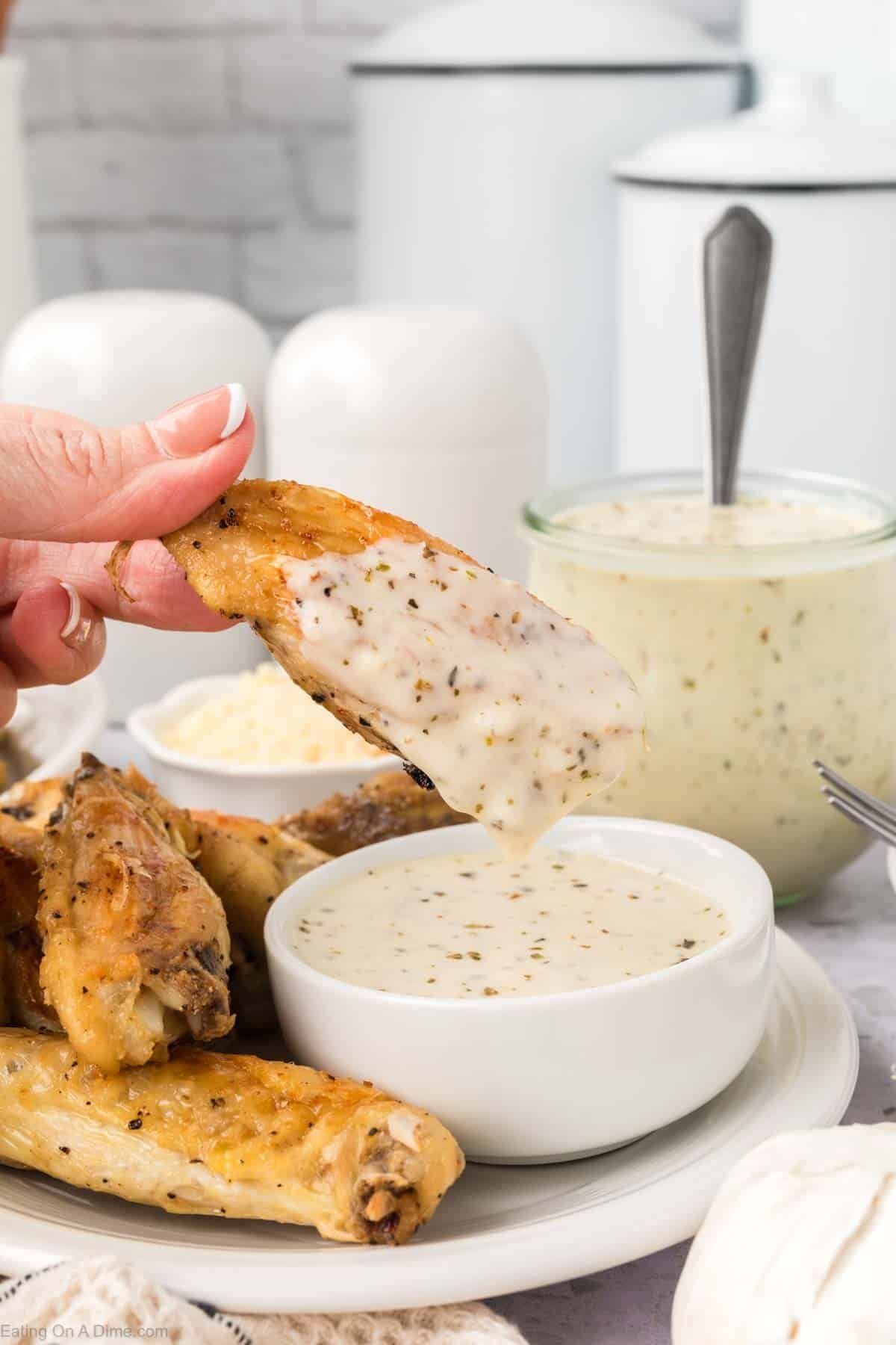 A hand dips a baked chicken wing into a bowl of creamy Garlic Parmesan Sauce, with more wings and a jar of the savory sauce in the background on a white table.