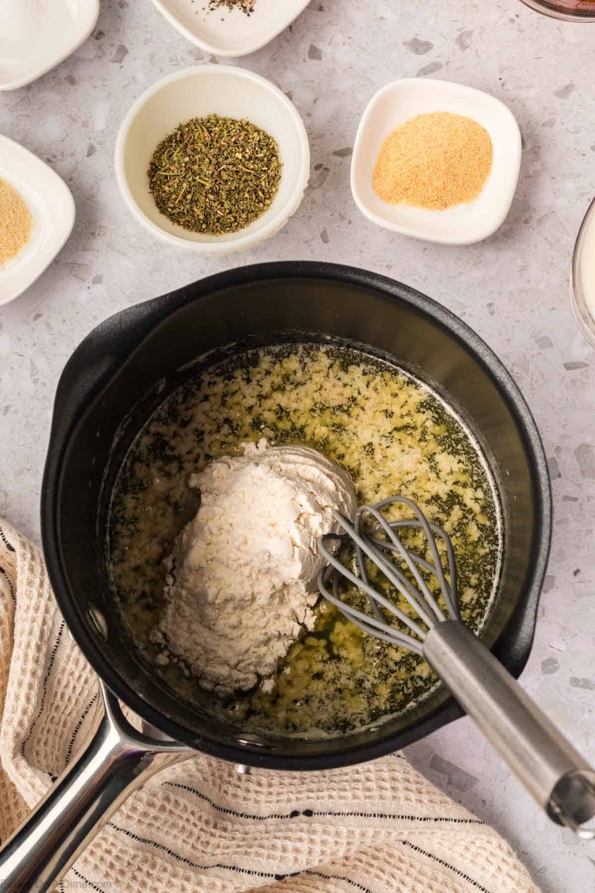 A saucepan with melted butter and flour being whisked together for a Garlic Parmesan Sauce, surrounded by small bowls of dried herbs, garlic powder, and pepper on a light countertop.