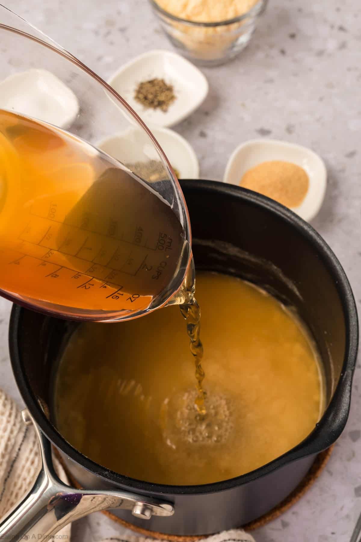 A measuring cup pours a golden-brown Garlic Parmesan Sauce into a saucepan with a similar liquid. In the background, small dishes hold seasoning and a bowl with grated cheese sit on the gray countertop.