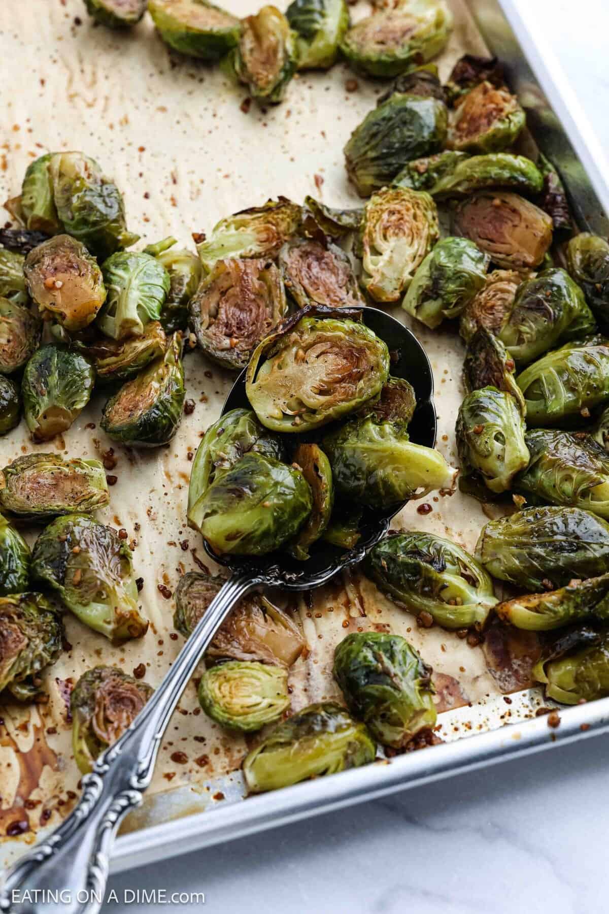 A baking sheet with Honey Balsamic Brussels Sprouts, some halved and browned, with a serving spoon lifting a portion. The sprouts appear crispy, seasoned, and glazed to perfection.