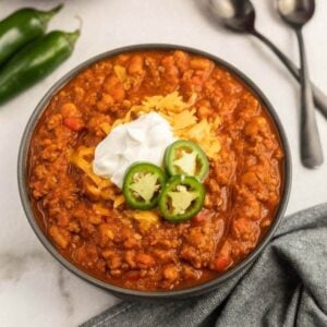 A bowl of chili from Meal Plan 76 is topped with shredded cheese, sour cream, and sliced jalapeños. It sits on a marble surface next to whole jalapeños, two spoons, and a gray cloth napkin.