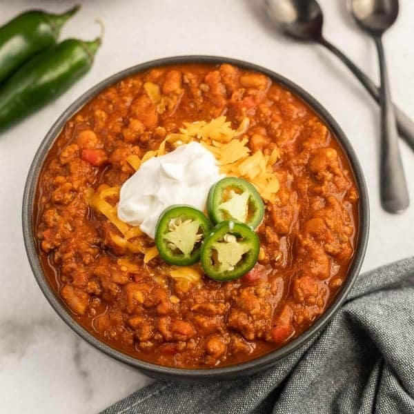 A bowl of chili from Meal Plan 76 is topped with shredded cheese, sour cream, and sliced jalapeños. It sits on a marble surface next to whole jalapeños, two spoons, and a gray cloth napkin.