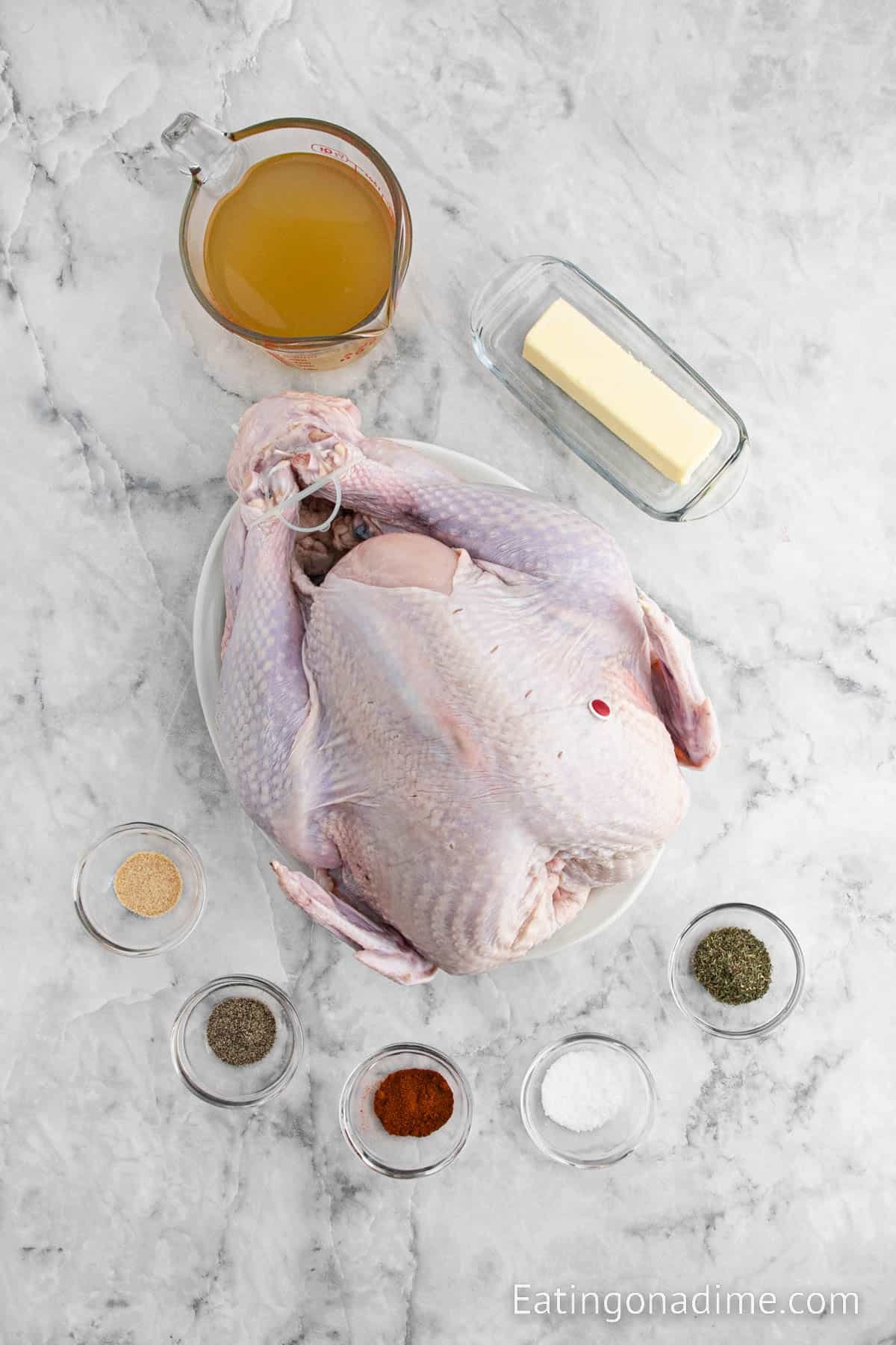 A raw whole turkey on a plate is surrounded by bowls of spices, a stick of butter in a dish, and a measuring cup of broth on a marble surface—perfect for preparing delicious grilled turkey. Text reads: Eatingonadime.com.
