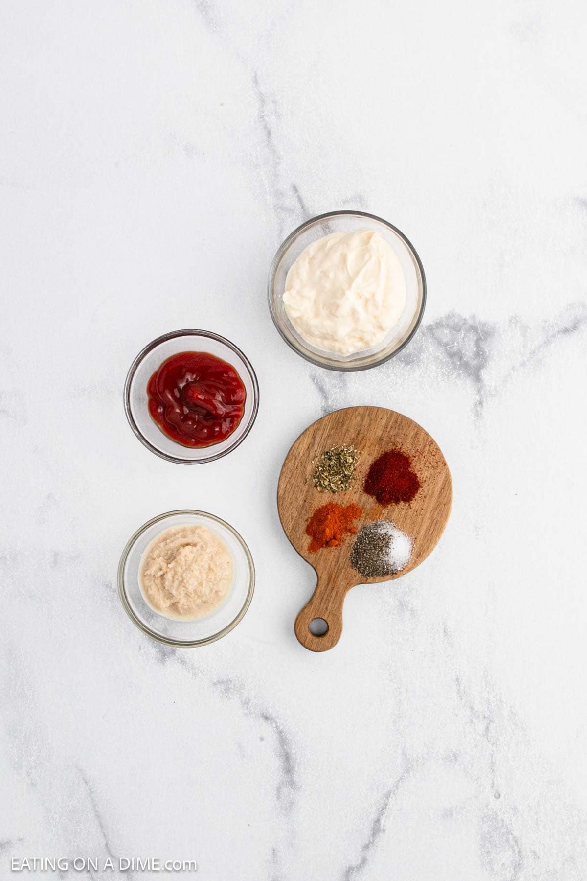 Three small bowls containing mayonnaise, ketchup, and horseradish sit next to a wooden board with piles of dried herbs and spices—perfect for dipping a crispy Bloomin Onion—on a white marble surface.