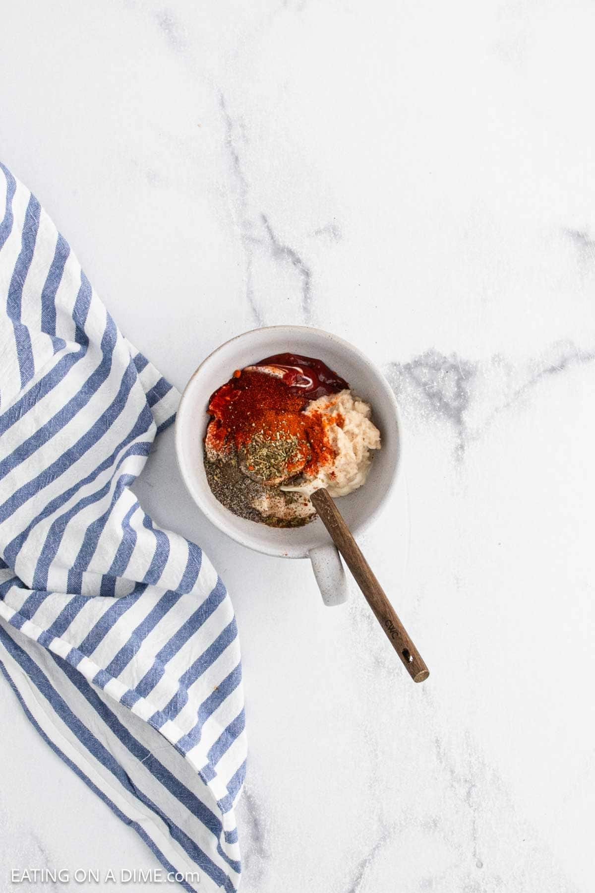A white bowl with a wooden spoon holds seasonings, ketchup, and mayonnaise—perfect for dipping your favorite bloomin onion. A blue and white striped cloth sits beside the bowl on a light marble surface.