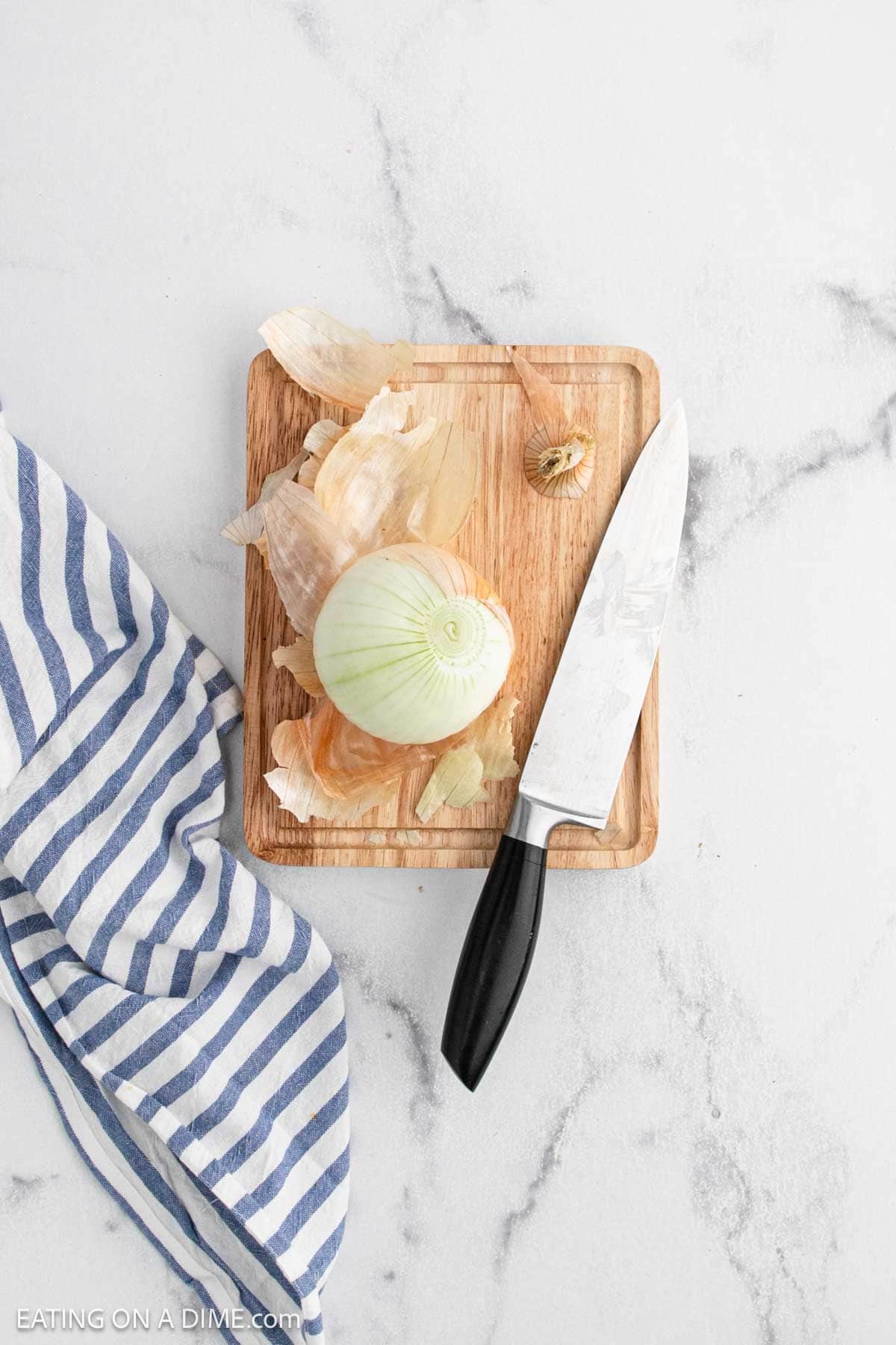 A half-peeled onion sits on a small wooden cutting board with a kitchen knife, ready to be transformed into a Bloomin Onion. Onion skins are scattered on the board, and a blue-and-white striped cloth rests nearby on the white marble surface.