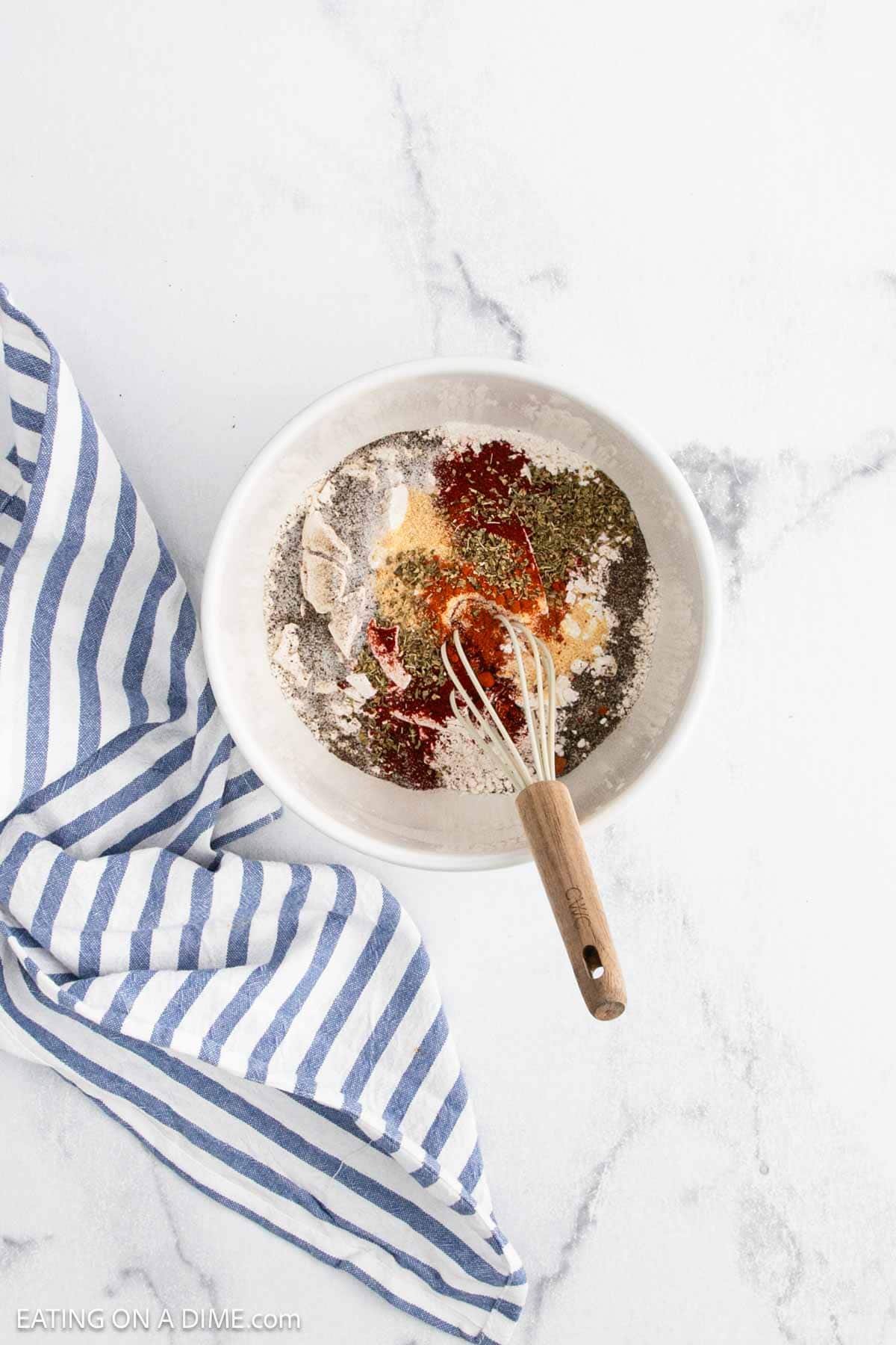 A white bowl filled with various colorful spices for making a Bloomin Onion and a whisk, placed on a white marble surface next to a blue and white striped kitchen towel.