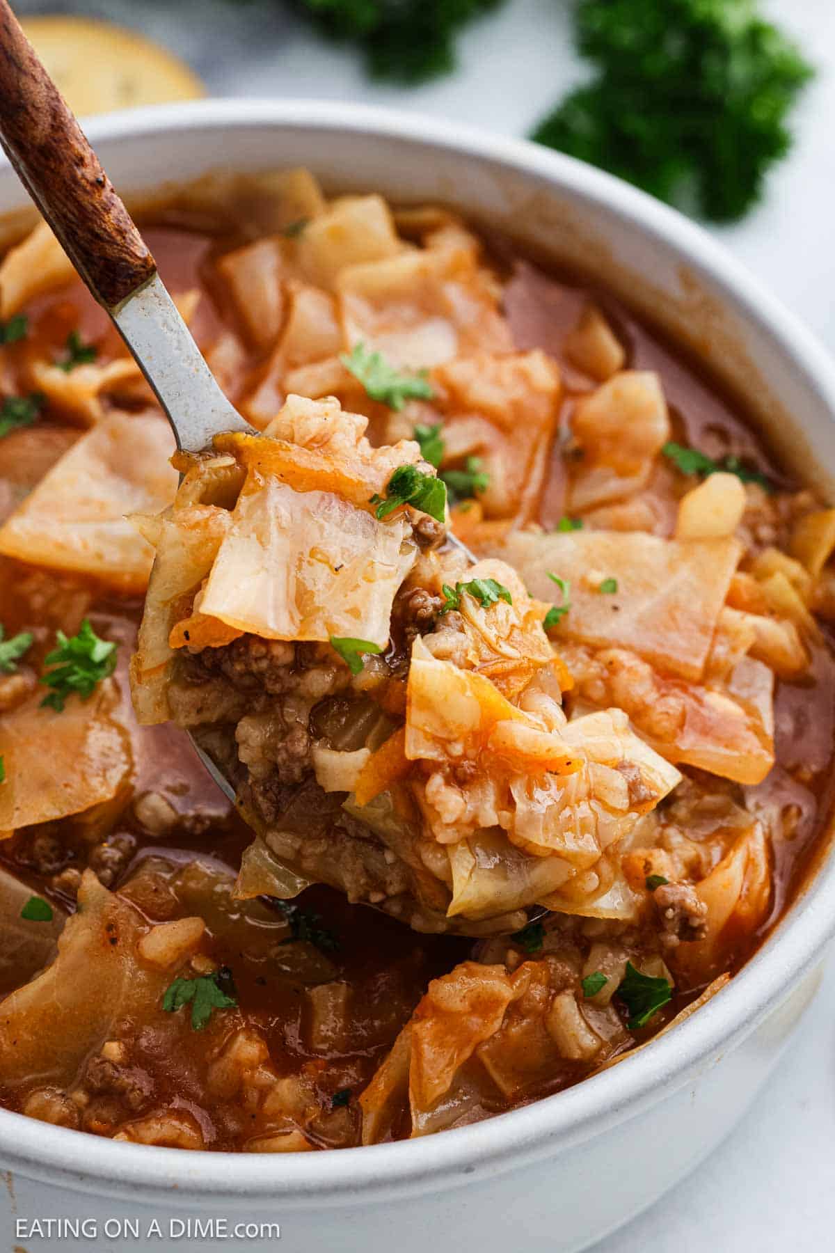 A close-up of a bowl of Stuffed Cabbage Soup with ground beef, cabbage, and rice in a tomato broth. A spoon is lifting a hearty portion from the bowl, and the soup is garnished with fresh chopped parsley.