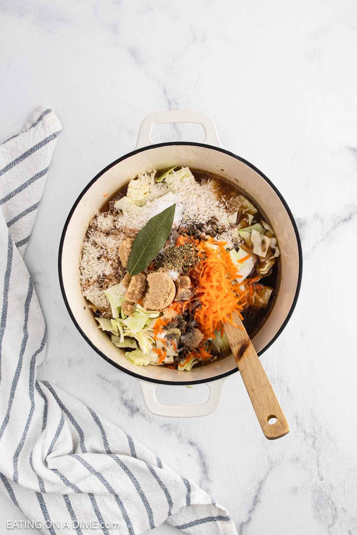 A white pot filled with ingredients for Cabbage Roll Soup, including shredded carrots, cabbage, spices, a bay leaf, and brown sugar, sits on a marble countertop beside a striped kitchen towel. A wooden spoon rests in the pot.