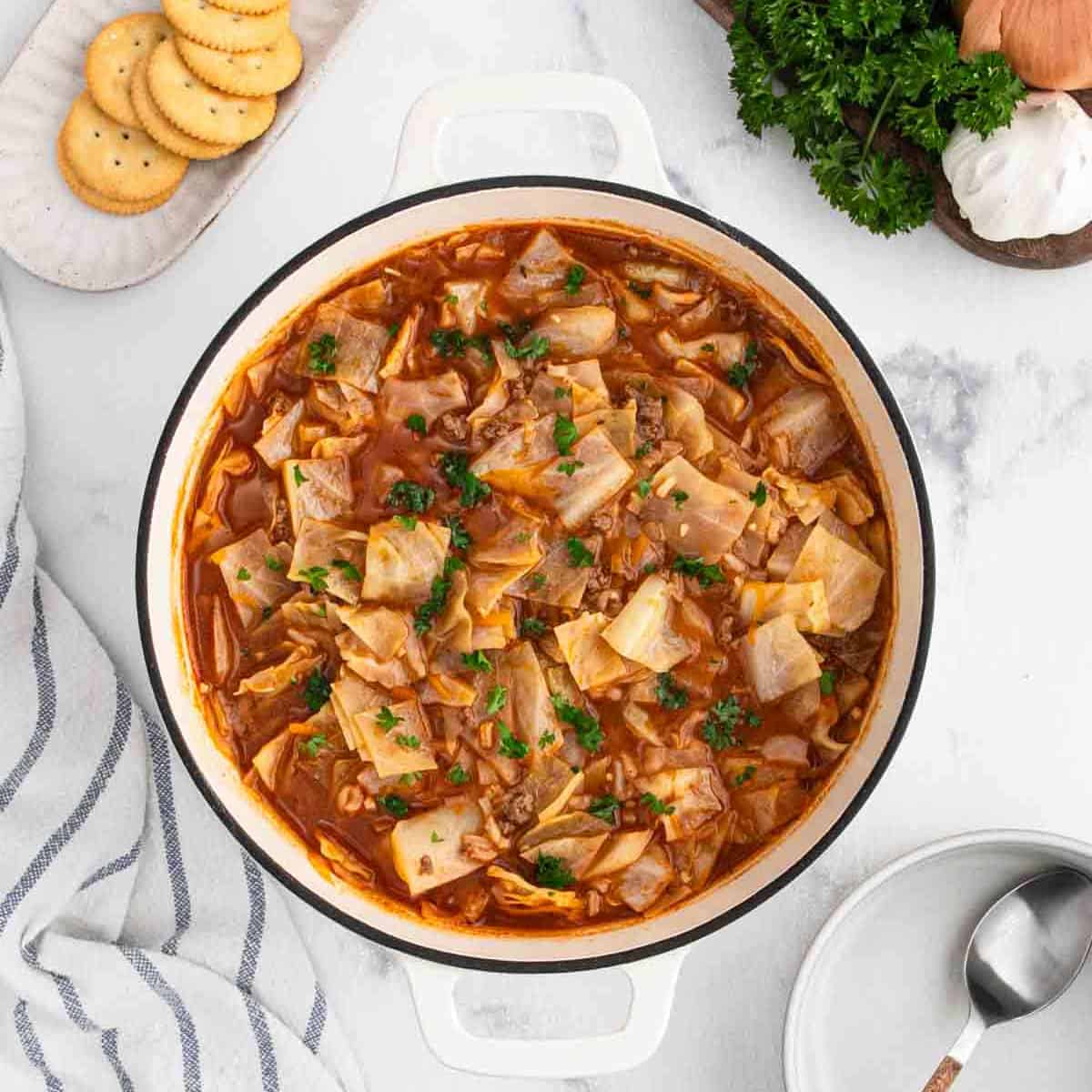 A white pot filled with hearty Cabbage Roll Soupโground meat, cabbage, and tomato broth, garnished with chopped parsley. Nearby are crackers, a napkin, spoon, and fresh parsley on a white marble surface.