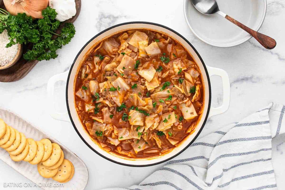 A pot of Cabbage Roll Soup garnished with herbs sits on a marble counter, surrounded by a bowl of crackers, a striped towel, a spoon, and a dish of chopped parsley.