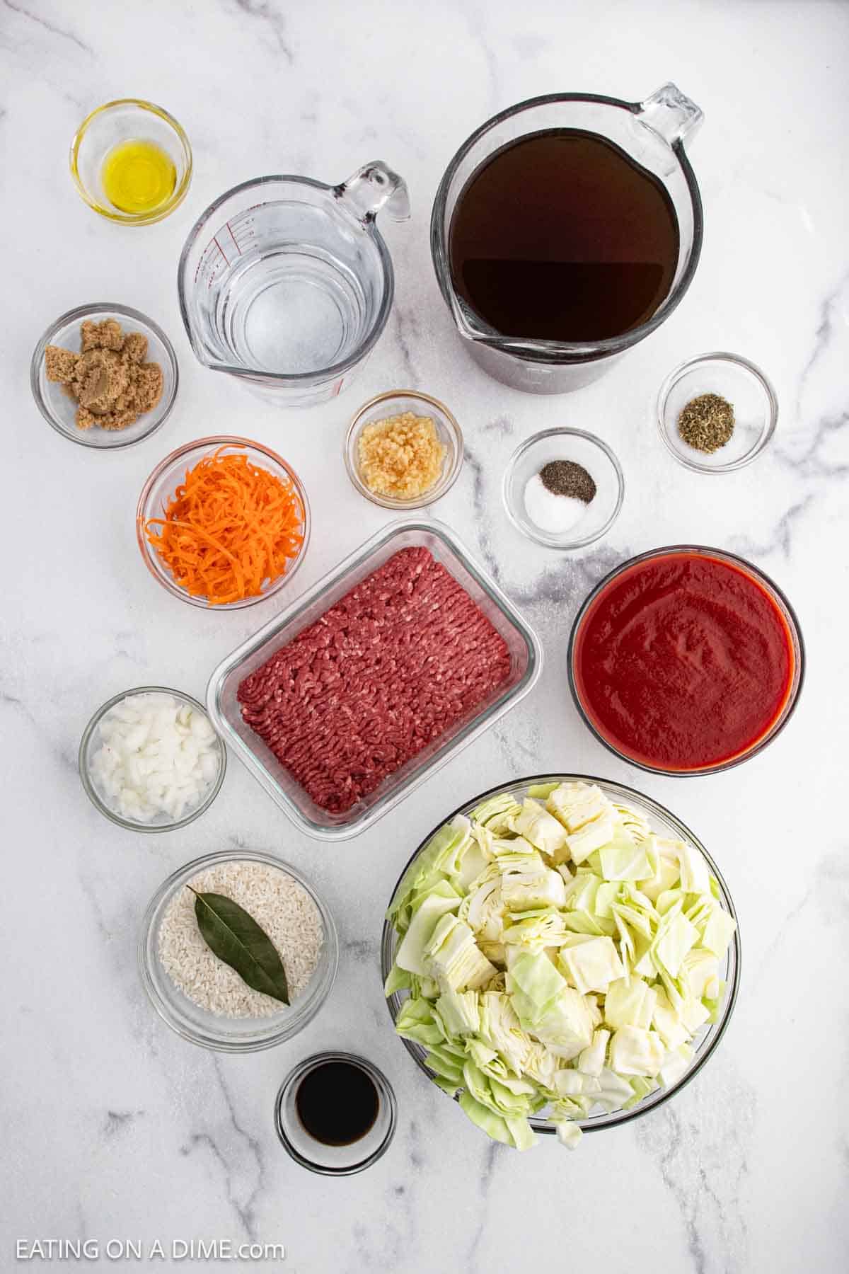 Top-down view of ingredients for Cabbage Roll Soup, featuring ground beef, chopped cabbage, carrots, diced onions, garlic, white rice, bay leaf, crushed tomatoes, broth, water, Worcestershire sauce, brown sugar, salt, and pepper.