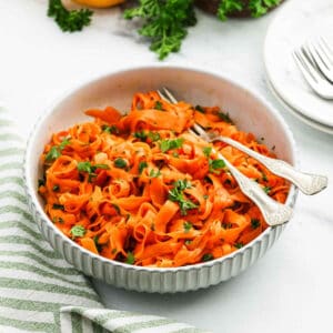 A white bowl filled with carrot salad ribbons garnished with chopped parsley. Two vintage-style forks rest in the bowl. The background includes a striped cloth, plates, and forks on a marble surface.