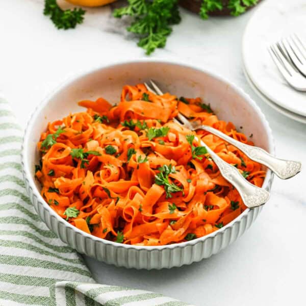 A white bowl filled with carrot salad ribbons garnished with chopped parsley. Two vintage-style forks rest in the bowl. The background includes a striped cloth, plates, and forks on a marble surface.