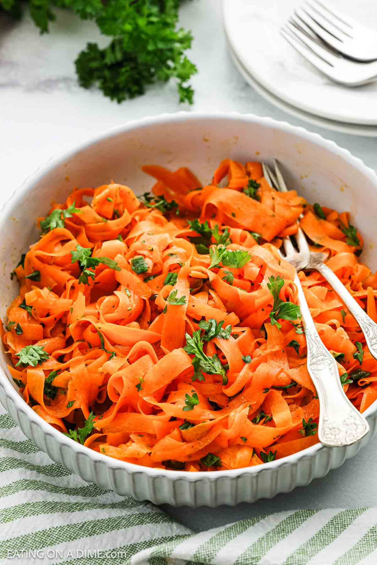 A white bowl filled with vibrant carrot salad, its ribbon-like strands garnished with fresh parsley. Two silver forks rest inside the bowl. Nearby, a stack of plates, cutlery, and a bunch of parsley complete the scene.