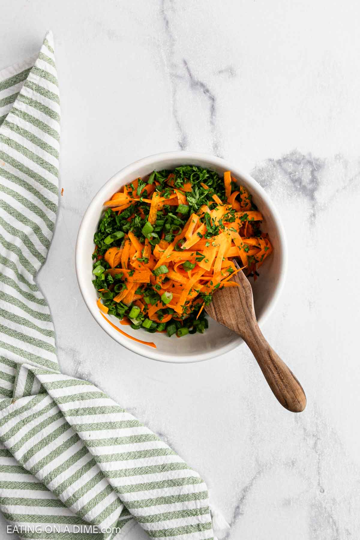 A white bowl filled with vibrant carrot salad—shredded carrots and chopped green herbs—rests on a marble surface beside a green and white striped kitchen towel, with a wooden spoon tucked inside.