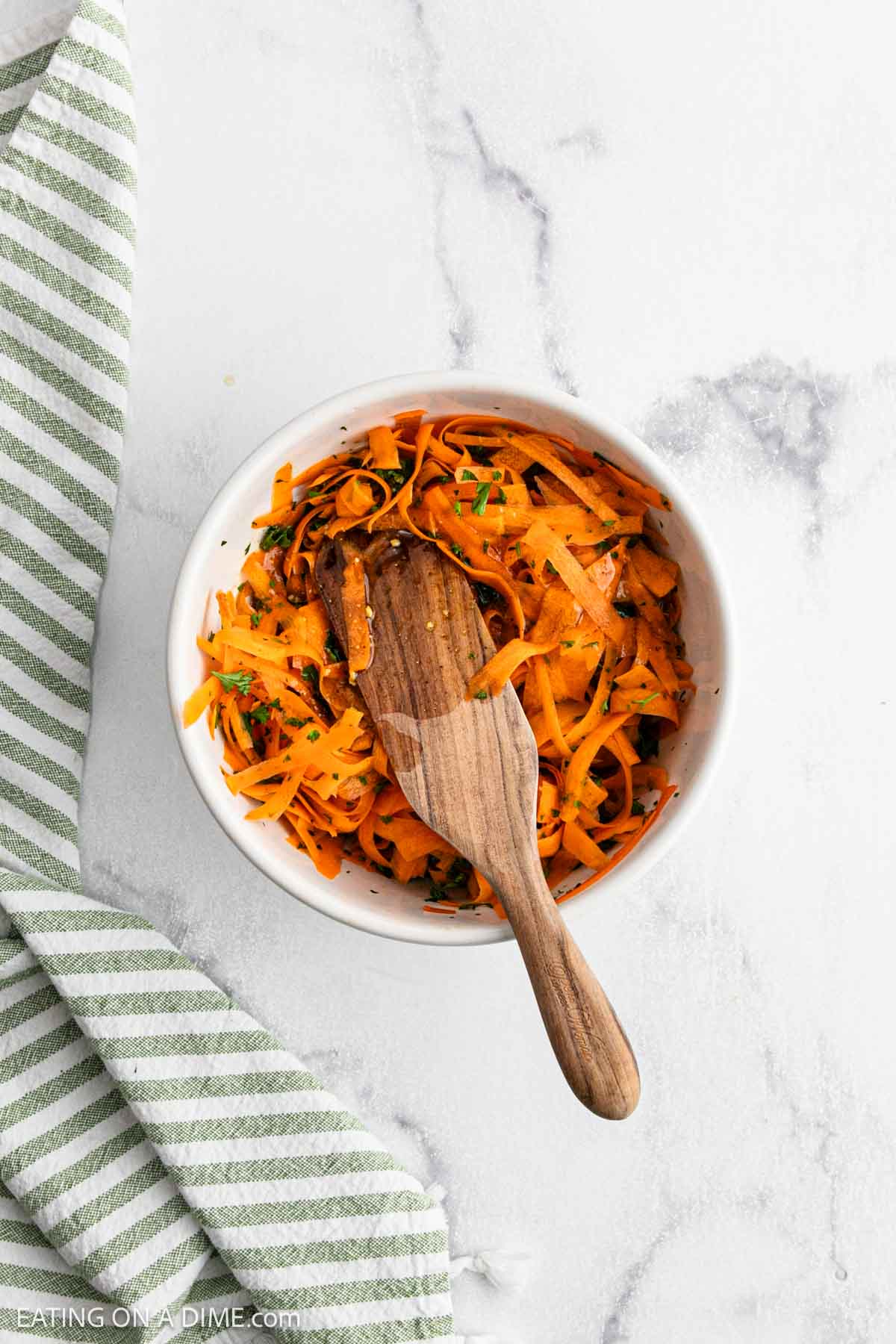 A white bowl filled with vibrant carrot salad and fresh herbs, with a wooden spoon resting inside. A green and white striped kitchen towel is placed beside the bowl on a marble surface.